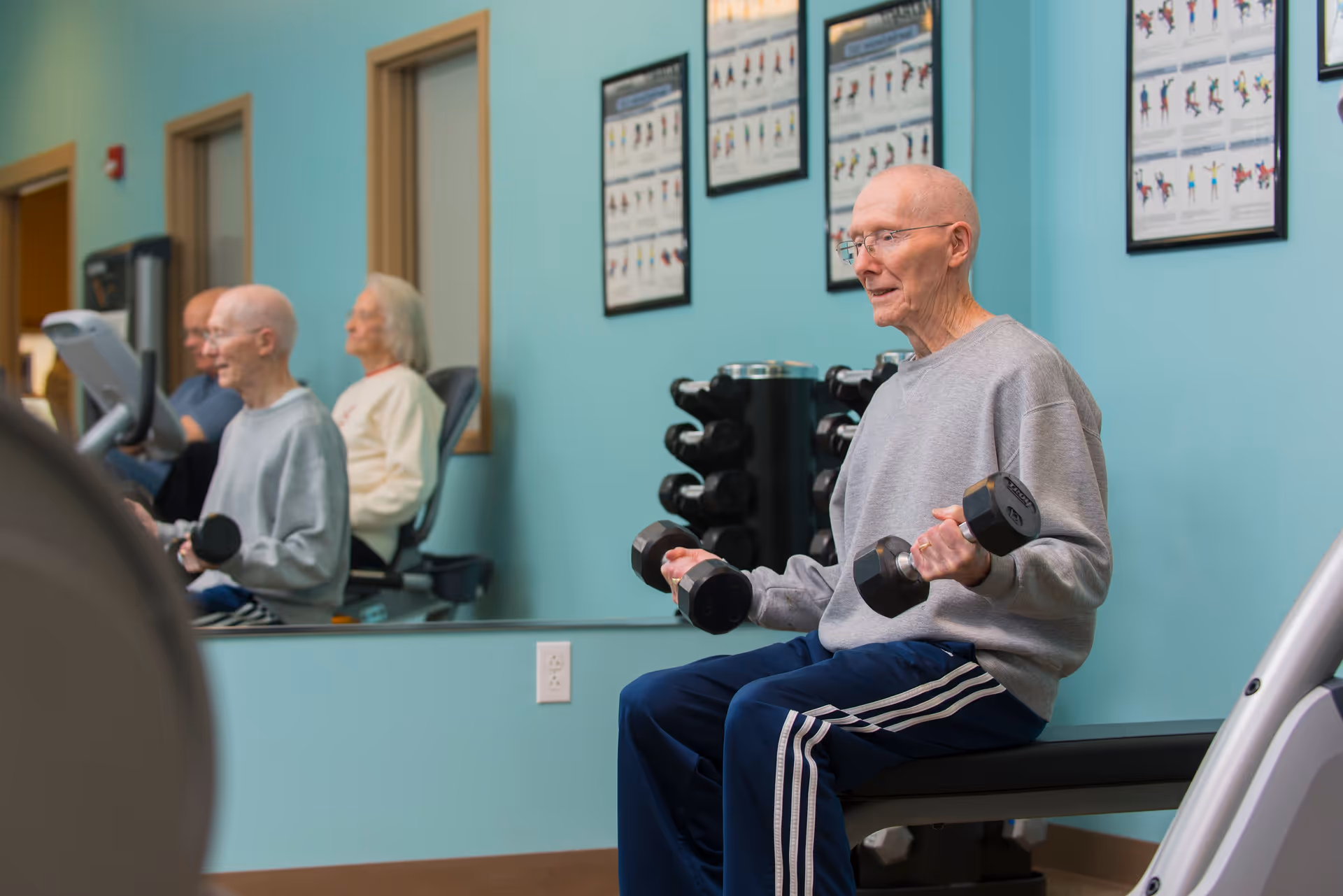 An elderly man wearing glasses, a gray sweatshirt, and blue track pants is sitting on a bench in a fitness room lifting dumbbells. Behind him is a mirror reflecting other seniors exercising on stationary bikes. The walls are painted light blue and have framed exercise instruction posters.