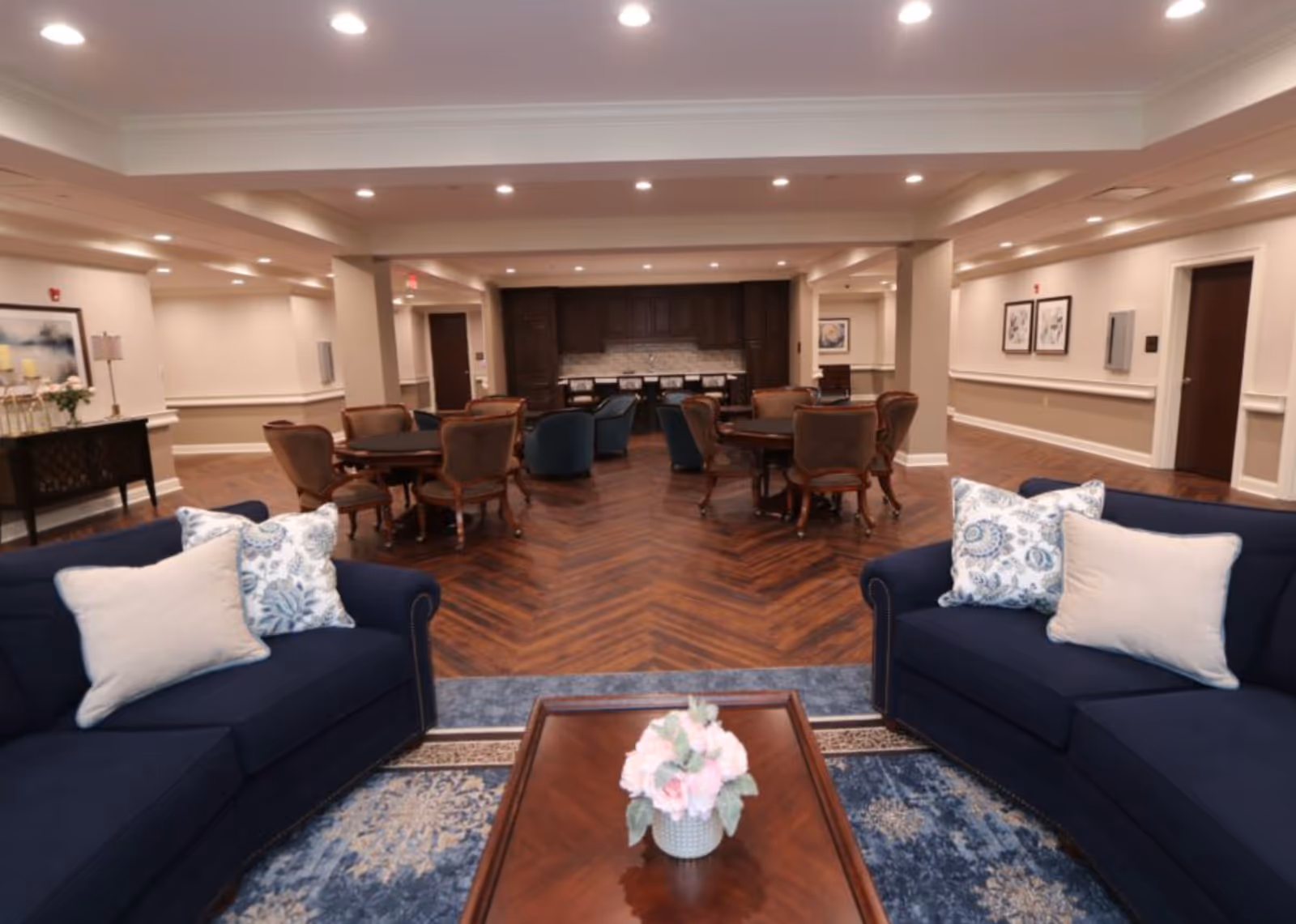 A spacious and well-lit common area in a senior living facility featuring two navy blue sofas with decorative pillows facing a wooden coffee table with a floral centerpiece. Behind the seating area, there are several round tables with chairs and a kitchenette with dark wood cabinets and a tiled backsplash. The room has wood-patterned flooring and neutral-colored walls with framed artwork.