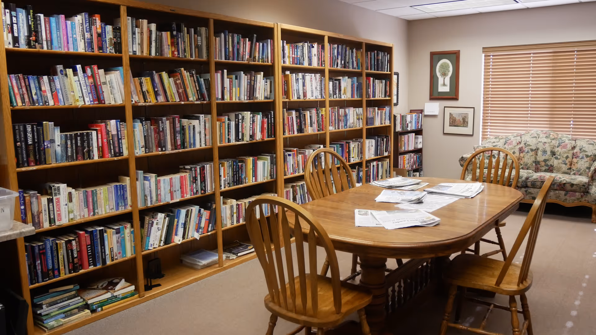 A cozy library room with wooden bookshelves filled with books along the wall, a wooden table with four wooden chairs around it, newspapers spread on the table, a floral patterned couch near a window with closed blinds, and framed artwork on the walls.