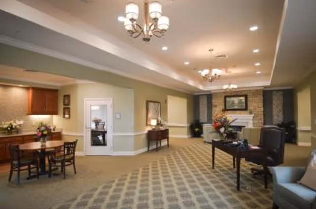 A spacious and well-lit living room area in The Ganzhorn Suites featuring patterned carpet flooring, multiple chandeliers, a fireplace with a framed picture above it, comfortable seating including armchairs and sofas, a wooden table with flowers, and a small dining table with chairs near a kitchenette area.