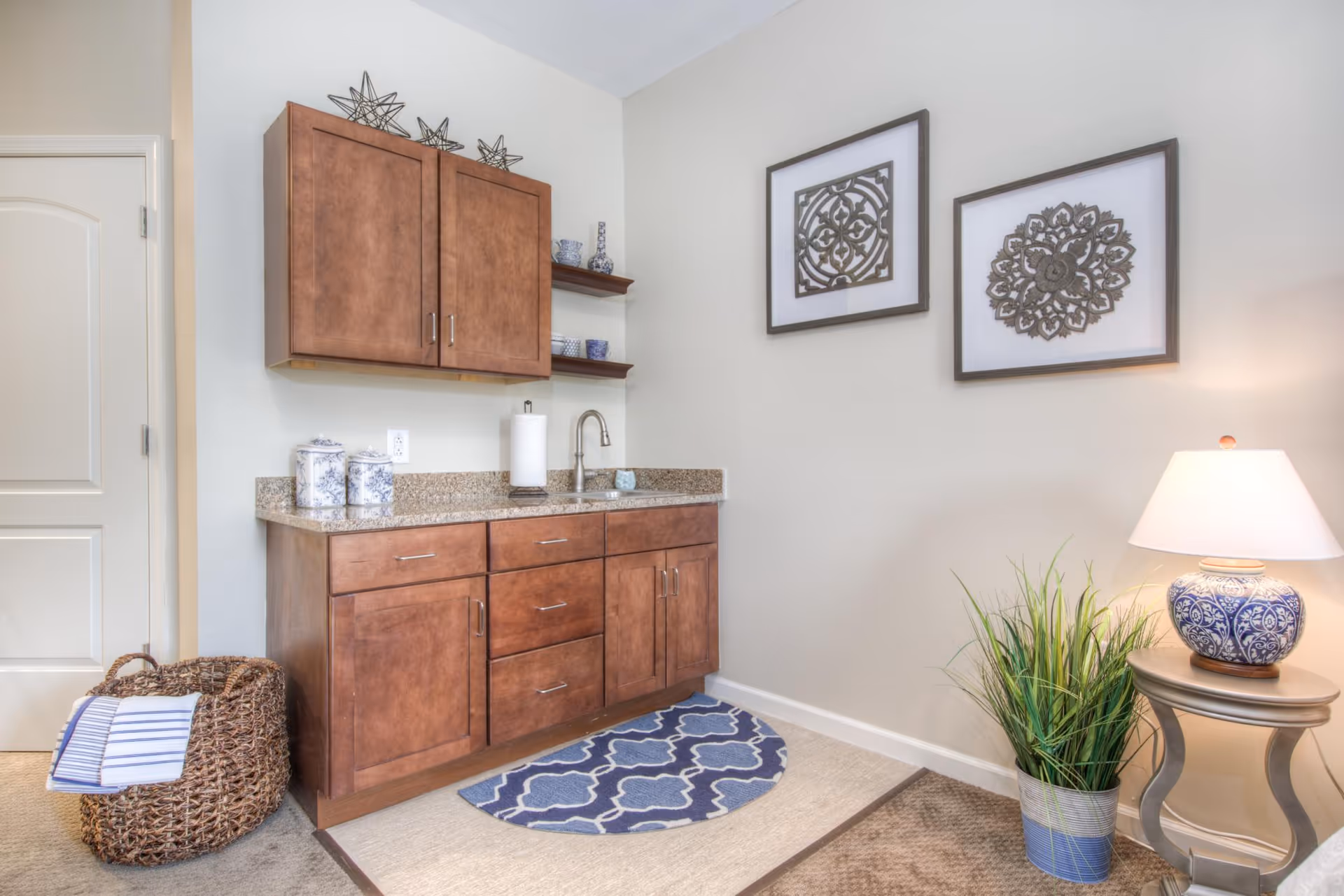 A small kitchenette area with wooden cabinets, a granite countertop, and a sink. There are decorative jars and a paper towel holder on the counter. Above the counter are two wooden shelves with blue and white dishes. On the wall are two framed decorative art pieces. To the right is a small round table with a blue and white lamp and a potted green plant. A woven basket with folded towels is on the floor to the left.