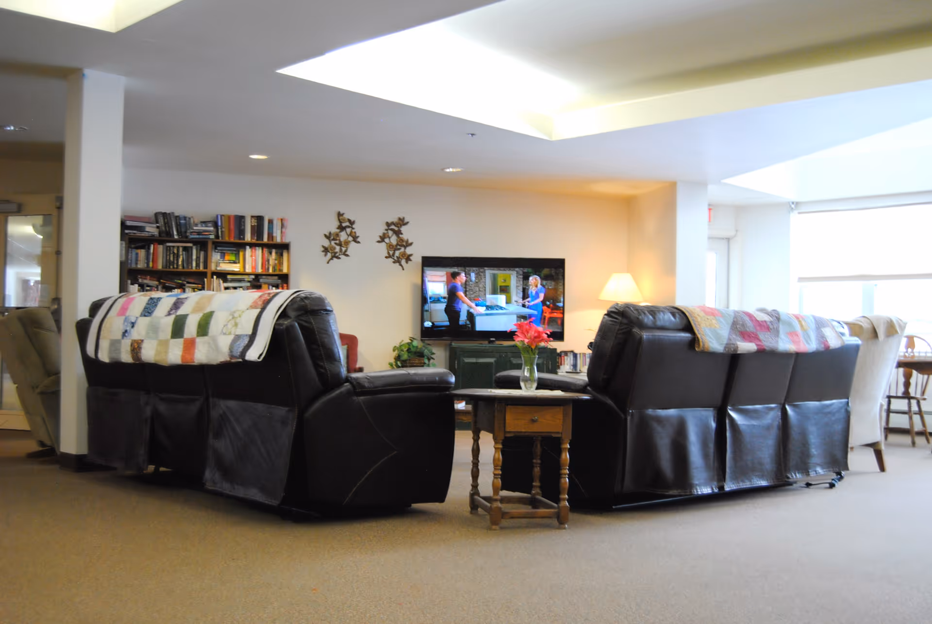 A cozy living room area with two black leather sofas covered with colorful patchwork quilts, facing a flat-screen TV mounted on the wall. A wooden side table with a vase of red flowers is placed between the sofas. In the background, there is a bookshelf filled with books and a lamp providing warm light. Large windows allow natural light to brighten the space.