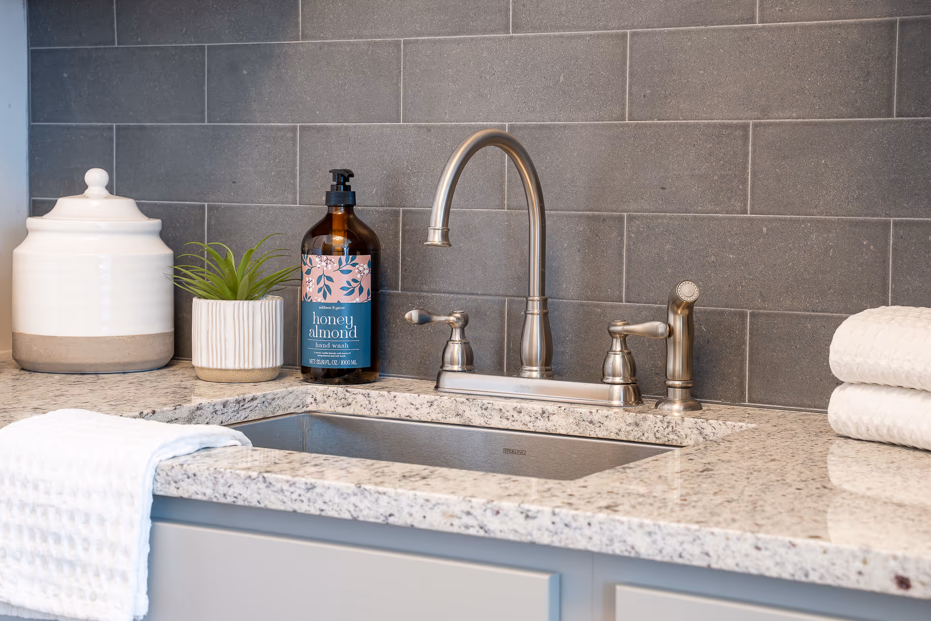 Close-up view of a kitchen sink with a modern silver faucet, a bottle of honey almond hand wash, a small potted plant, a white ceramic jar, and folded white towels on a granite countertop with gray tiled backsplash.