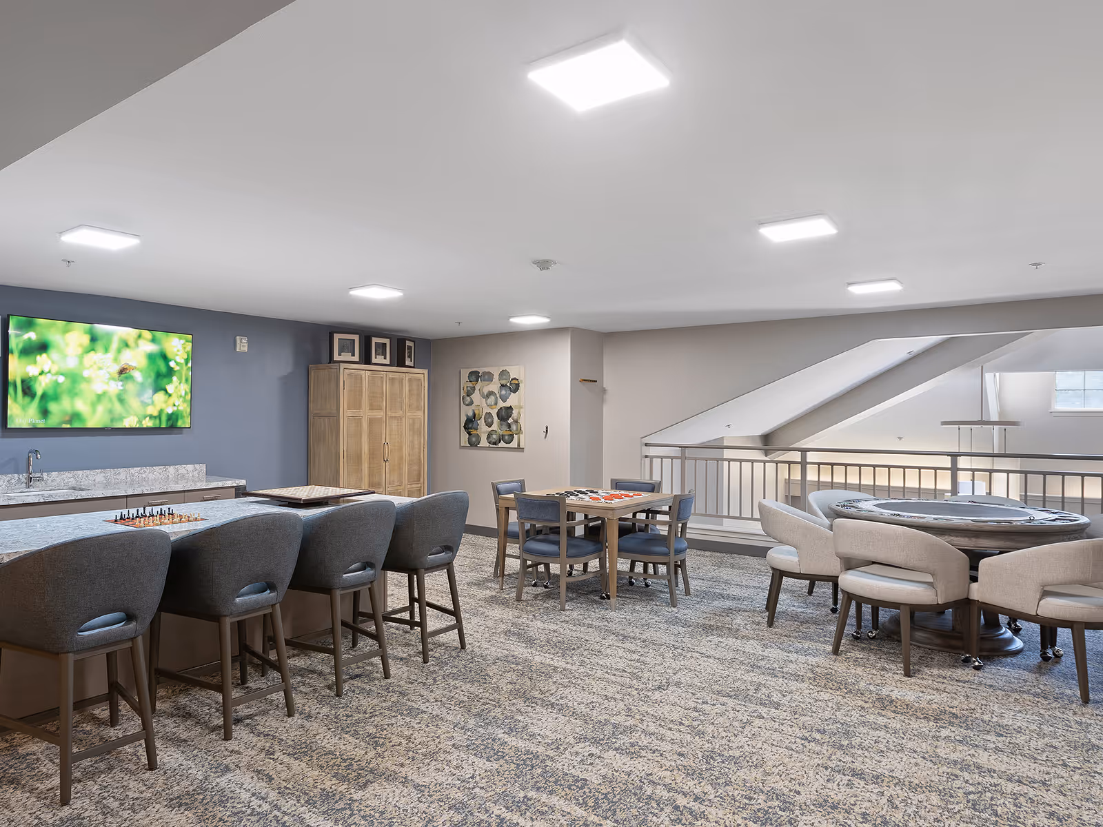 A spacious, well-lit common area with a long counter and four gray upholstered bar stools. The counter has a chessboard and another game board on it. There are two smaller tables with chairs, each set up with board games. A large TV is mounted on the blue accent wall above a countertop with a sink. The room has carpeted flooring and modern ceiling lights, with a railing overlooking a lower level.