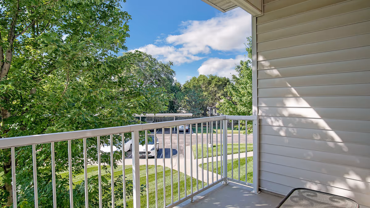 View from a balcony with white railing overlooking a parking area with cars, green grass, and trees under a partly cloudy blue sky.