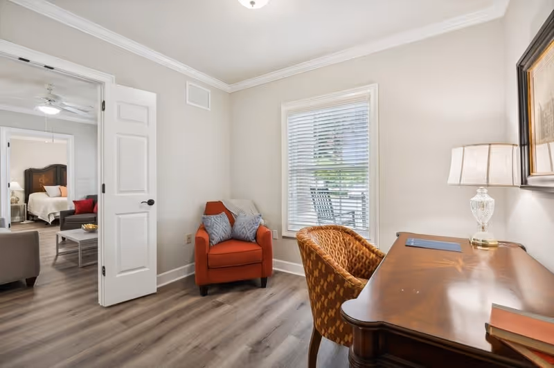 A cozy interior room with a wooden desk and a patterned chair next to a window with blinds. A table lamp and a book are on the desk. An orange armchair with two gray pillows is placed against the wall. Through an open door, a bedroom with a bed, nightstand, and ceiling fan is visible.