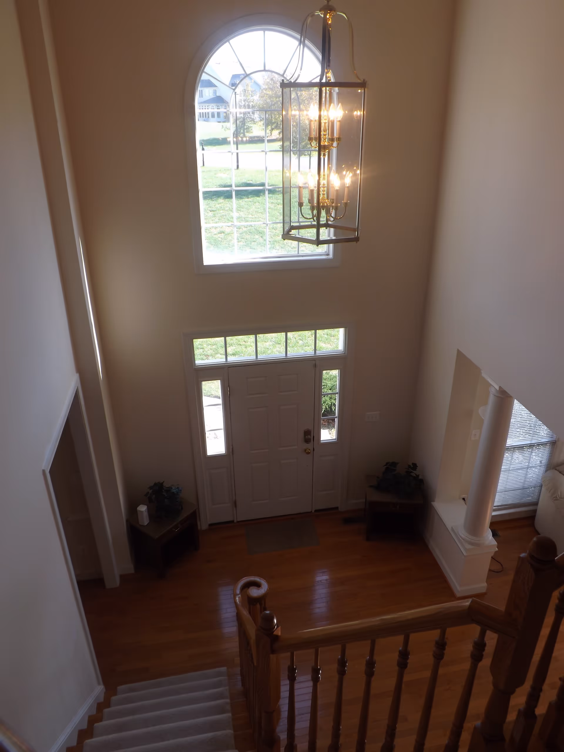 View from the top of a staircase looking down into a foyer with a white front door, sidelights, and a large arched window above. There is a hanging chandelier light fixture, hardwood floors, two small tables with plants on either side of the door, and a partial view of a living room area with a column and window blinds.