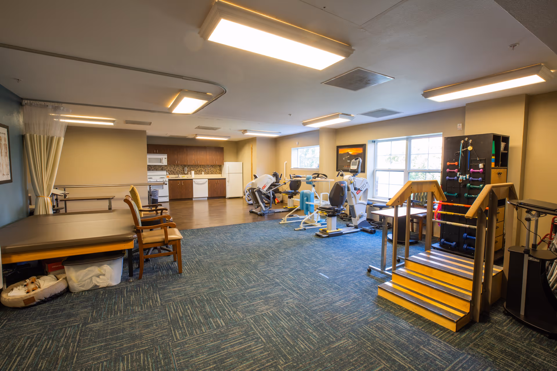 A senior living facility therapy and exercise room with exercise machines, parallel bars, practice stairs and a kitchenette in the background.