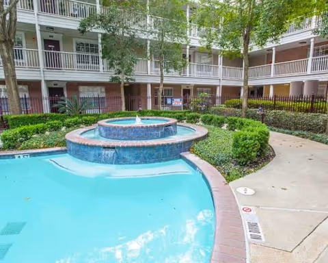 Outdoor courtyard area of a residential facility featuring a circular tiered water fountain surrounded by a small pool, landscaped greenery, trees, and a multi-story building with balconies in the background.