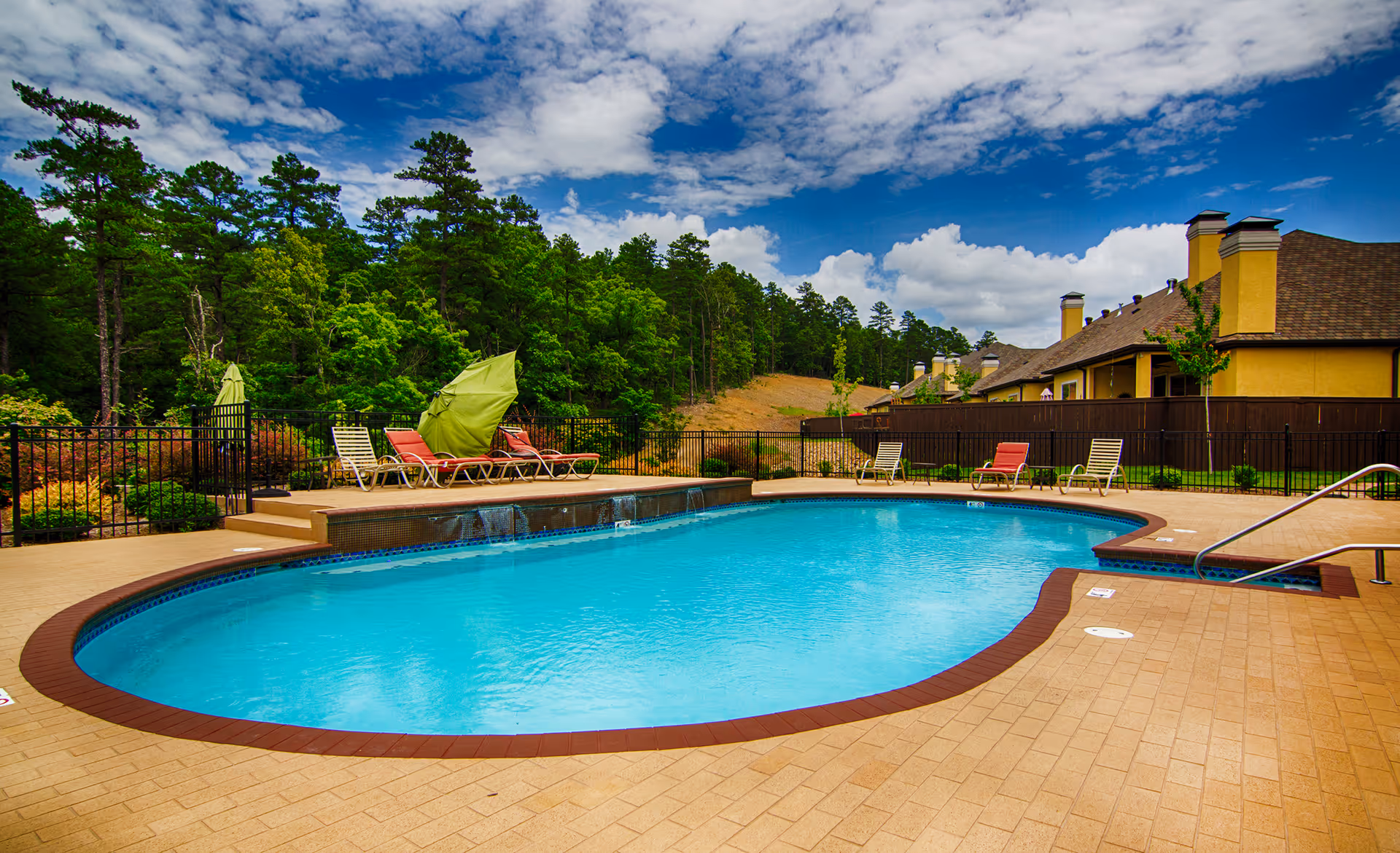 Outdoor swimming pool with lounge chairs and umbrellas next to townhome buildings and trees under a partly cloudy sky.