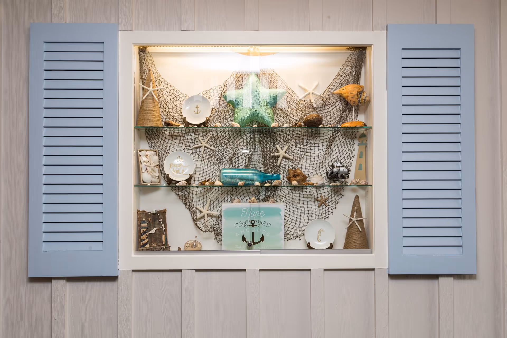 A decorative wall display featuring two light blue shutters on either side of a recessed glass shelf. The shelves hold various nautical-themed items including starfish, a green starfish sculpture, a fishing net backdrop, seashells, a blue glass bottle, small plates with anchor designs, a lighthouse figurine, and a sign with the word 'Hope' and an anchor illustration.