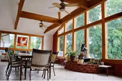 Sunlit dining/sitting area with large floor-to-ceiling windows, exposed wooden beams, tile floor, a table with chairs, and potted plants.