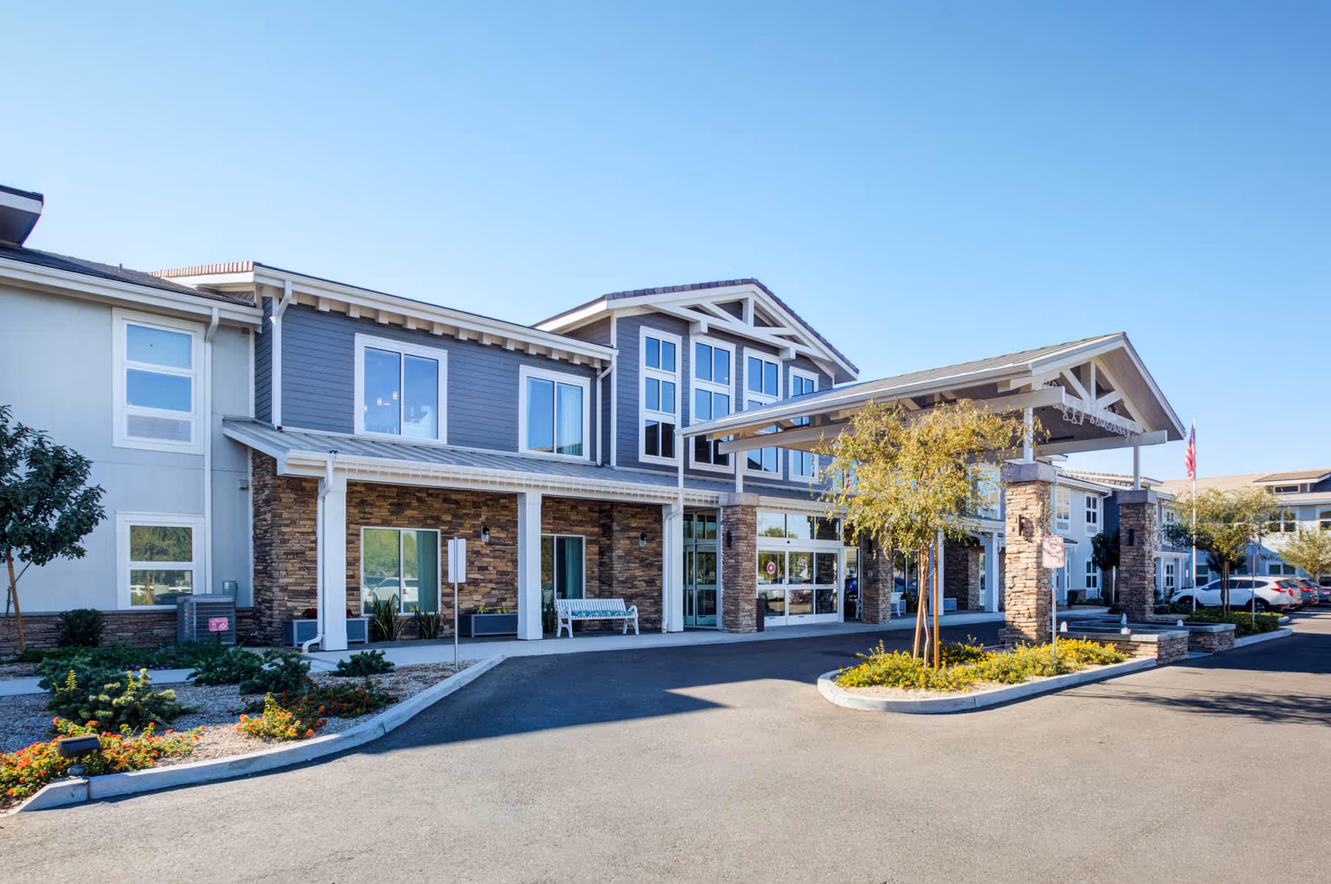 Exterior view of a senior living facility building with a covered entrance supported by stone pillars, landscaped greenery, and a clear blue sky.