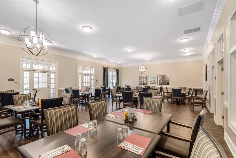 A spacious dining room with multiple wooden tables and chairs arranged neatly. Each table has placemats, glasses, and silverware set up. The room has large windows and glass doors letting in natural light, beige walls, and modern light fixtures hanging from the ceiling. There are framed pictures on the far wall and curtains on one window.