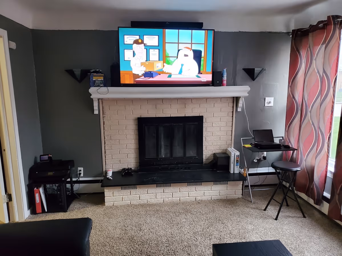 A living room with a beige brick fireplace and a black screen mounted above it displaying a cartoon show. The room has gray walls, a carpeted floor, and a window with red and beige patterned curtains on the right. There is a small black table with a laptop and a stool near the window, and a printer on a stand to the left of the fireplace.