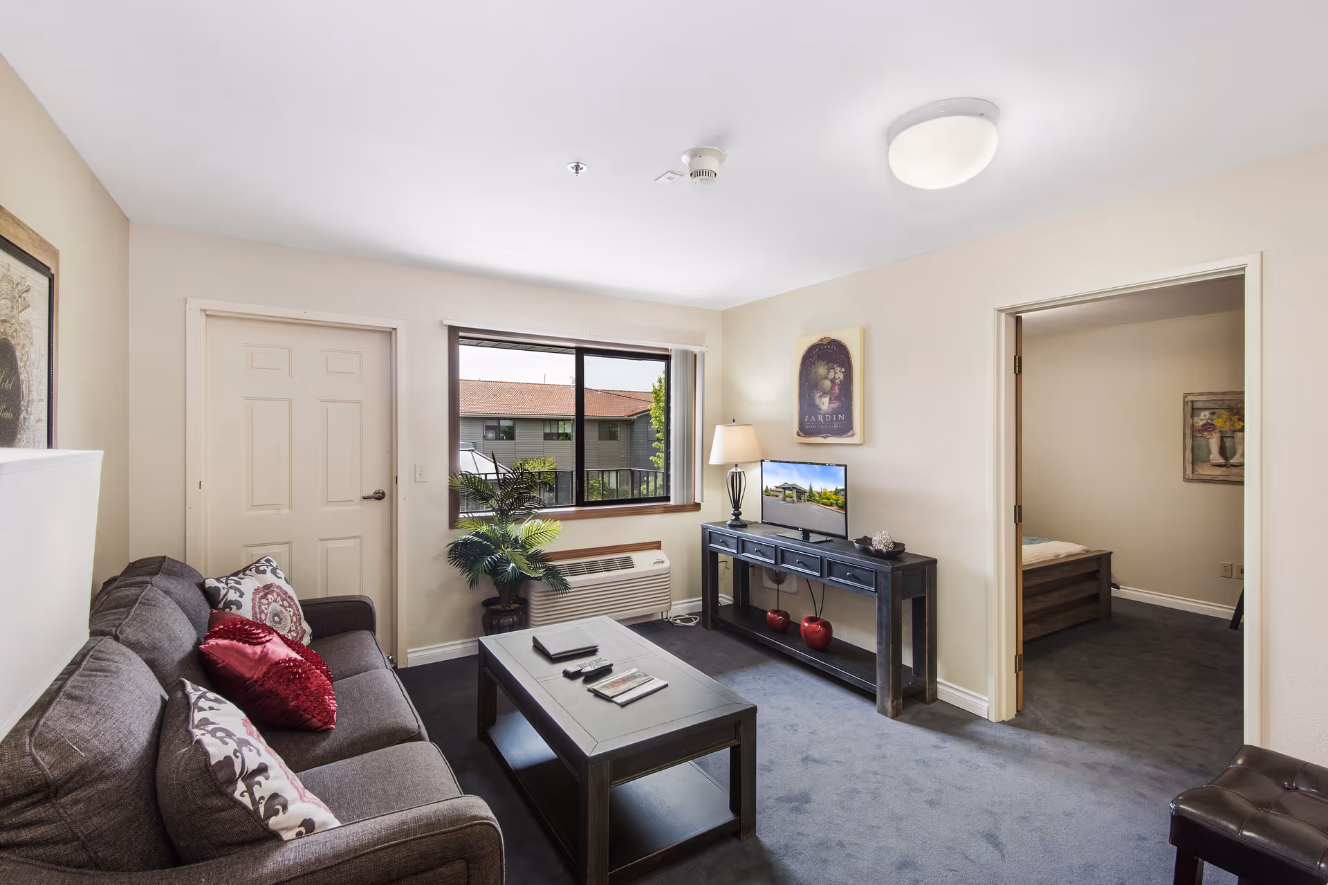 A cozy living room in a retirement and assisted living facility featuring a gray sofa with decorative pillows, a black coffee table with magazines and remote controls, a black console table with a lamp and a small TV, a potted plant near the window, and an open doorway leading to a bedroom with a bed and wall art.
