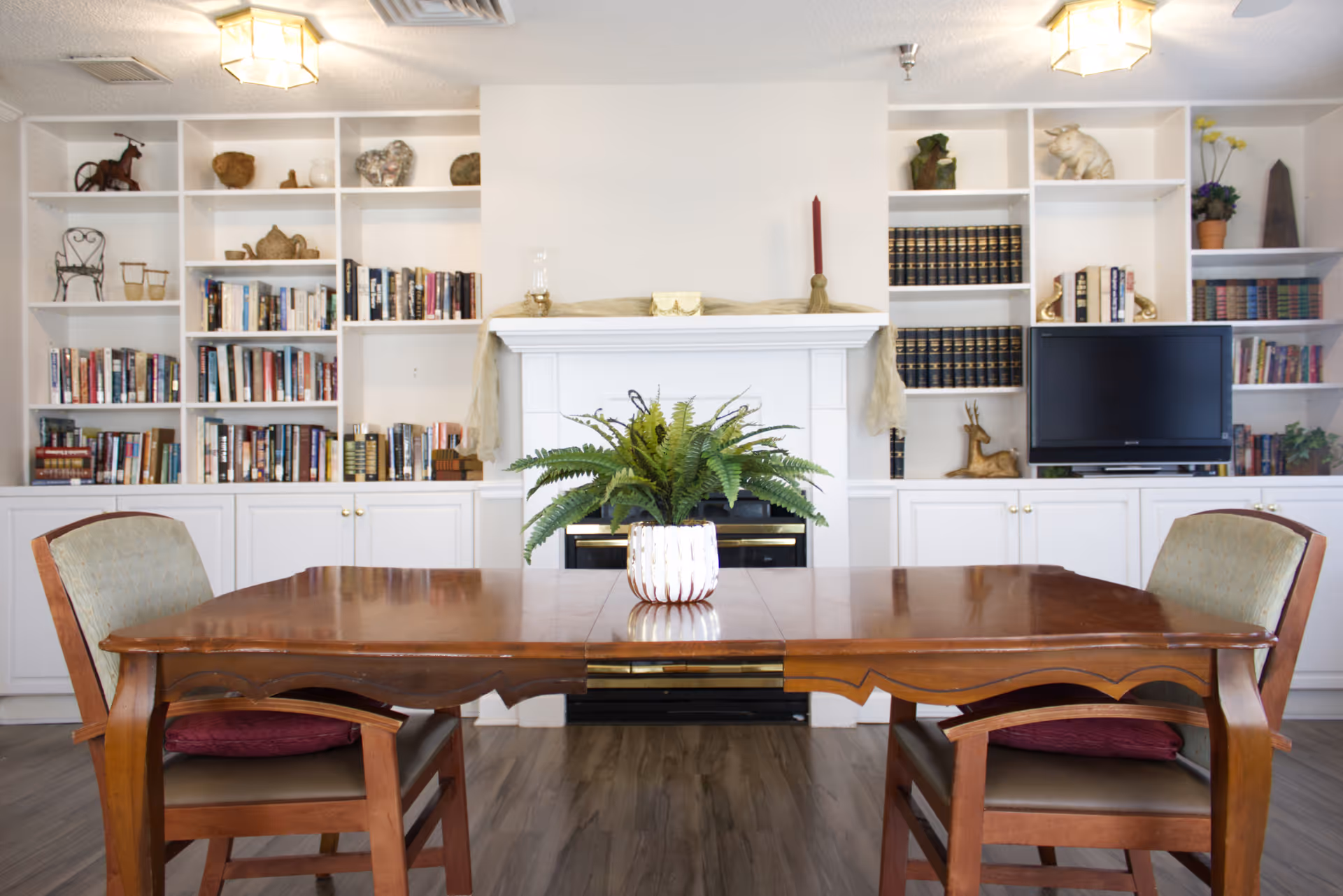 A cozy interior room featuring a wooden table with two cushioned chairs, a potted fern centerpiece on the table, a white fireplace in the background, and built-in white bookshelves filled with books, decorative items, and a television.