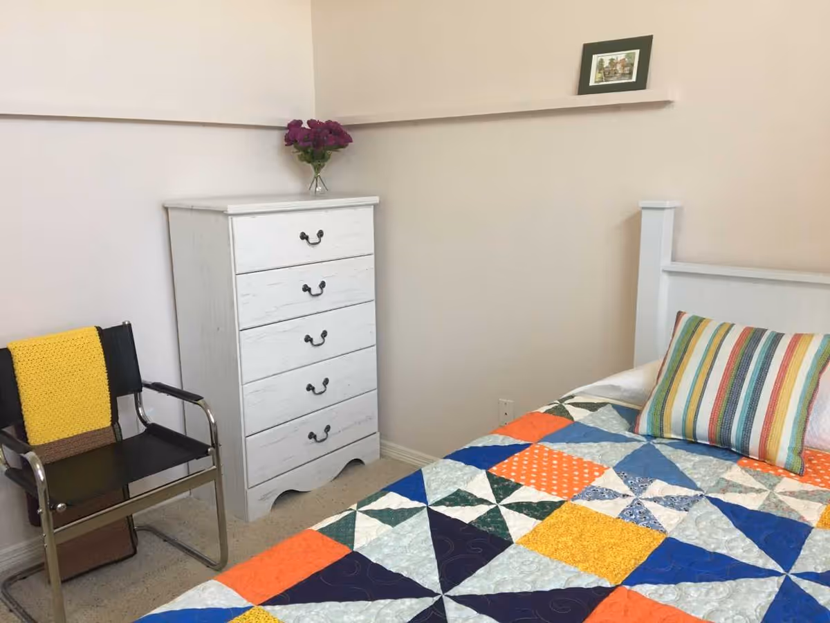 A bedroom corner with a white wooden dresser topped with a vase of purple flowers, a black chair with a yellow knitted blanket draped over it, and a bed with a colorful patchwork quilt and a striped pillow. A small framed picture is placed on a narrow wall shelf above the dresser.