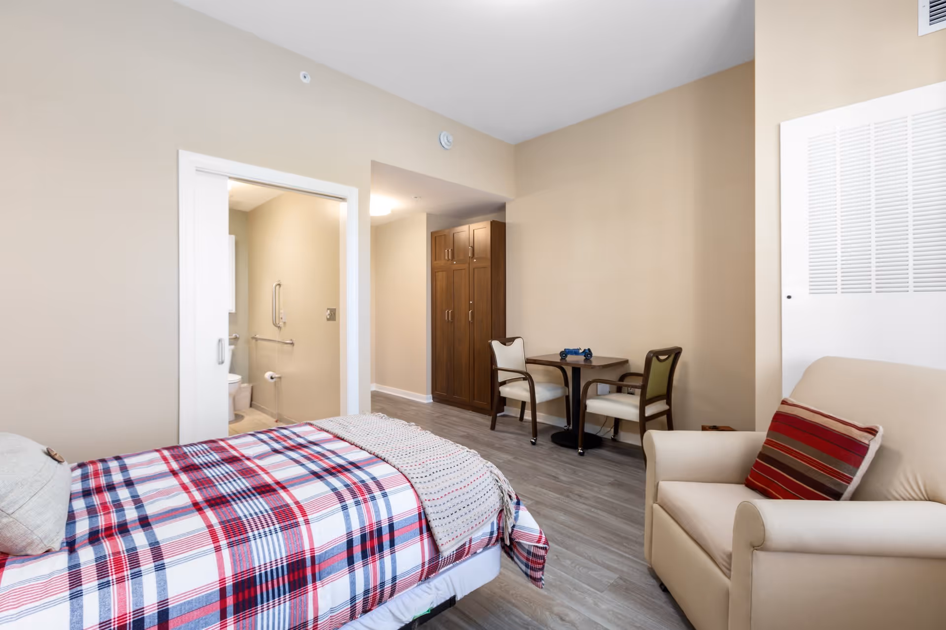 A cozy senior living bedroom with a bed covered in a red, white, and blue plaid blanket, a beige armchair with a striped pillow, a small table with two chairs, and a wooden wardrobe. An open door reveals a bathroom with grab bars and a toilet.