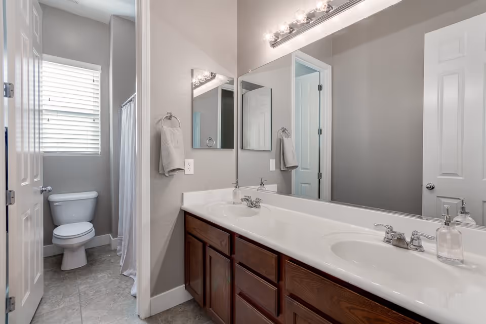 A clean bathroom with a double sink vanity featuring wooden cabinets and a large mirror above. To the left, there is a separate area with a toilet and a window with blinds, next to a shower with a white curtain.