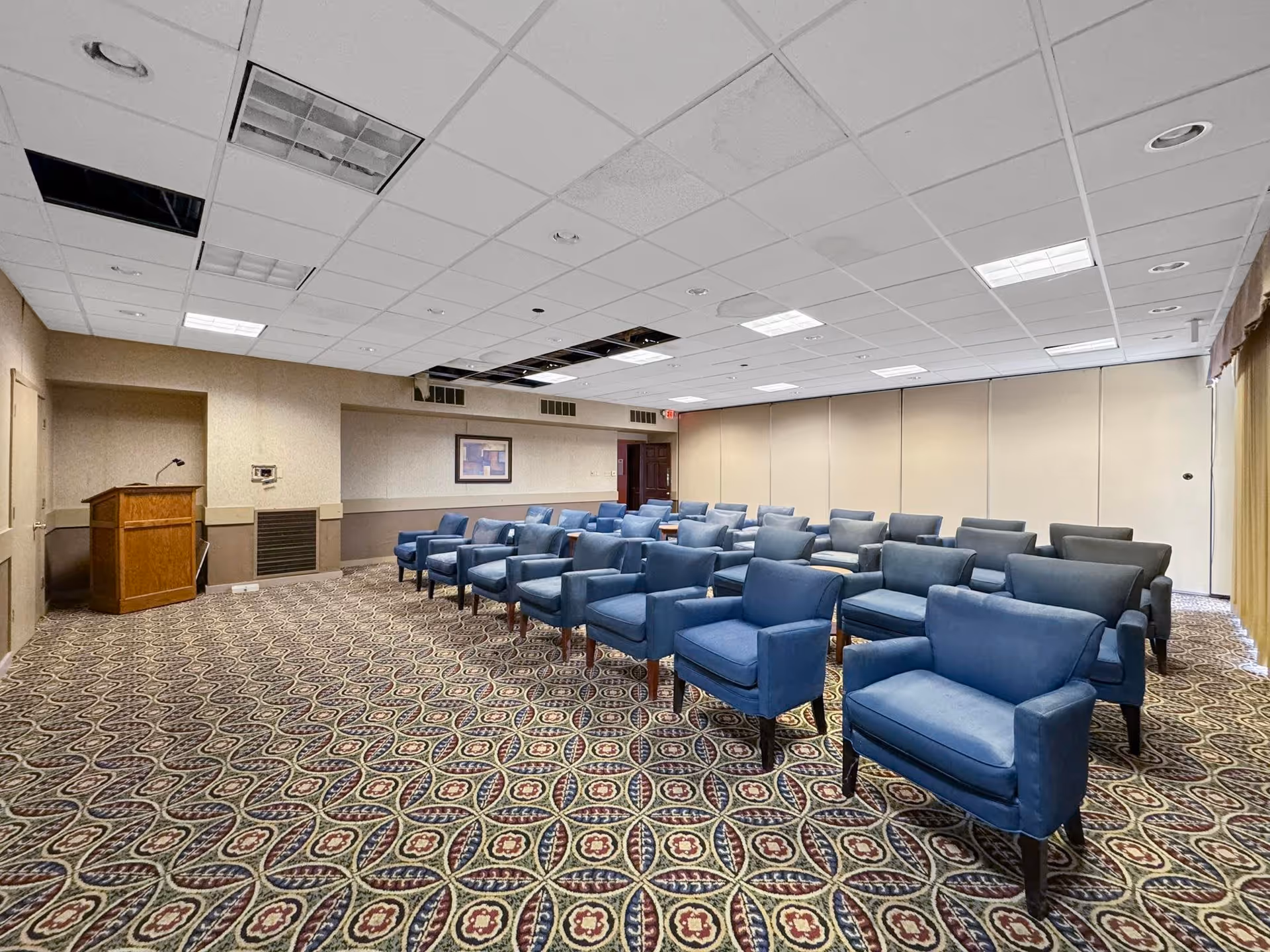 A carpeted meeting room with rows of blue upholstered armchairs arranged facing a wooden podium under a tiled ceiling.