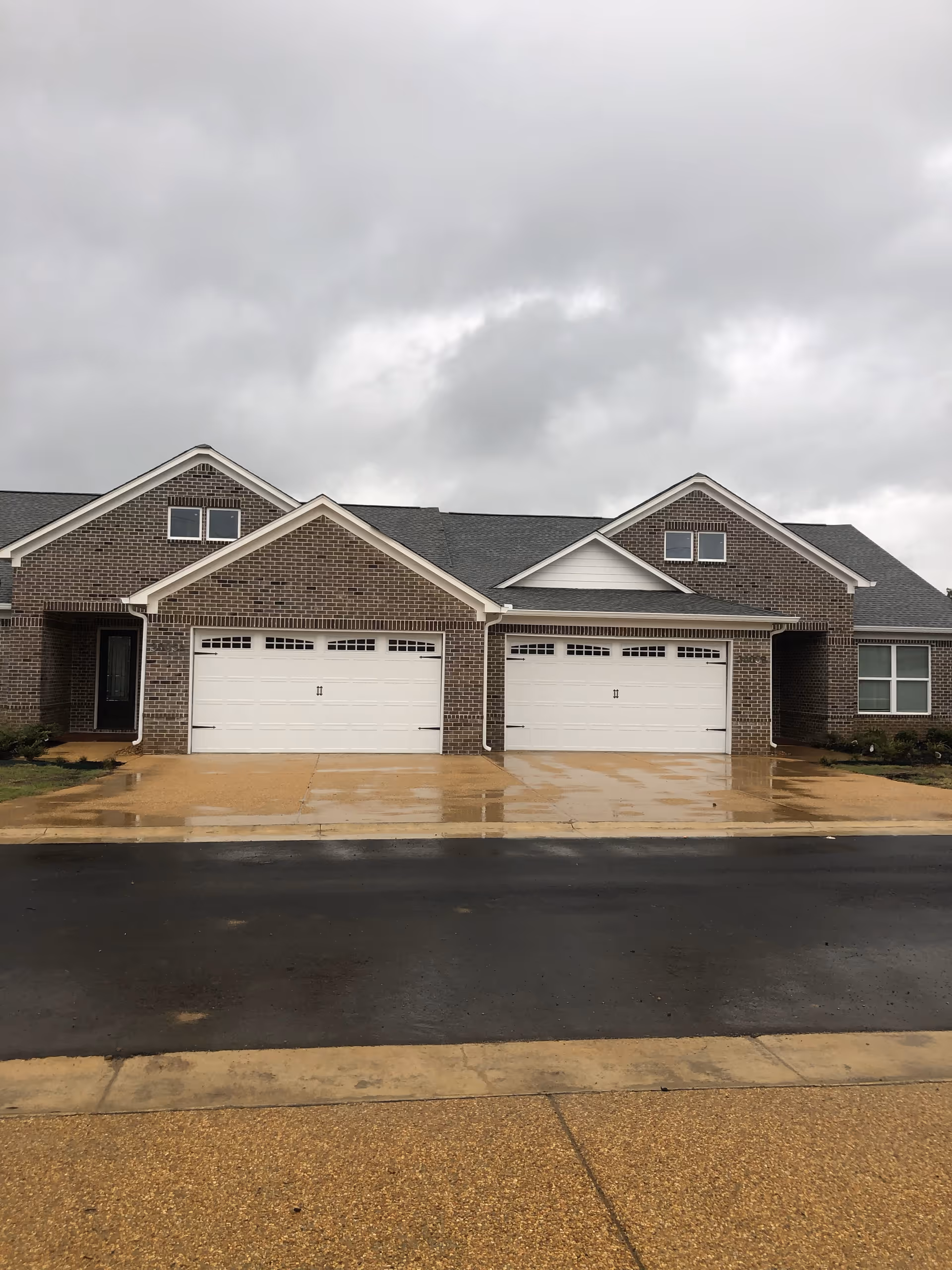 Exterior view of a brick residential building with two white garage doors and a wet driveway under a cloudy sky.