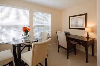 Bright dining nook with a glass-top table, upholstered chairs, a vase of orange flowers, and a writing desk with lamp in front of window blinds.