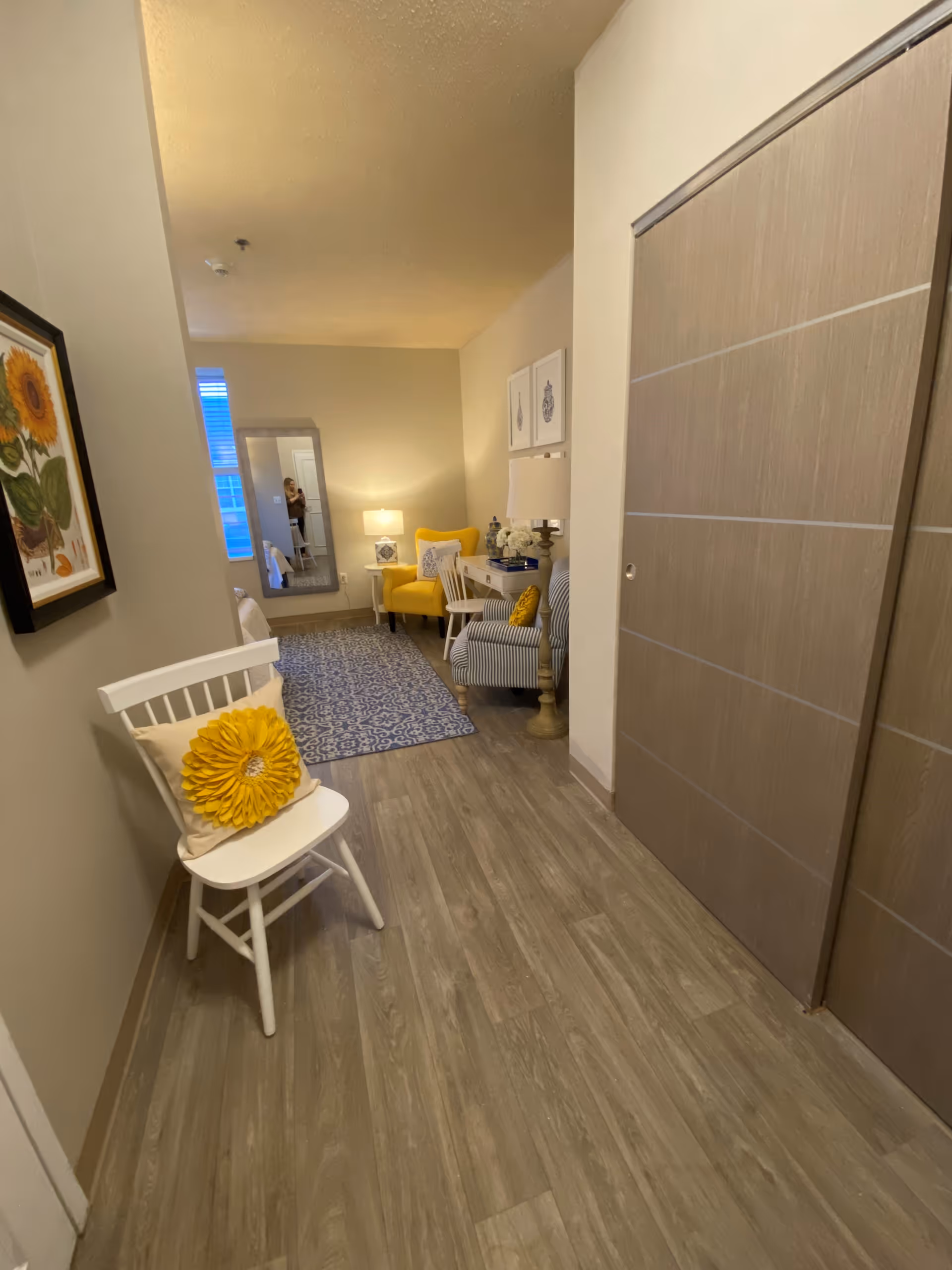 A hallway leading to a cozy sitting area with a yellow armchair, a striped armchair, a small white desk with a lamp, and decorative items. There is a white chair with a yellow flower cushion in the foreground, a blue patterned rug on the floor, and framed artwork on the walls. A large mirror reflects part of the room and a window with blinds.