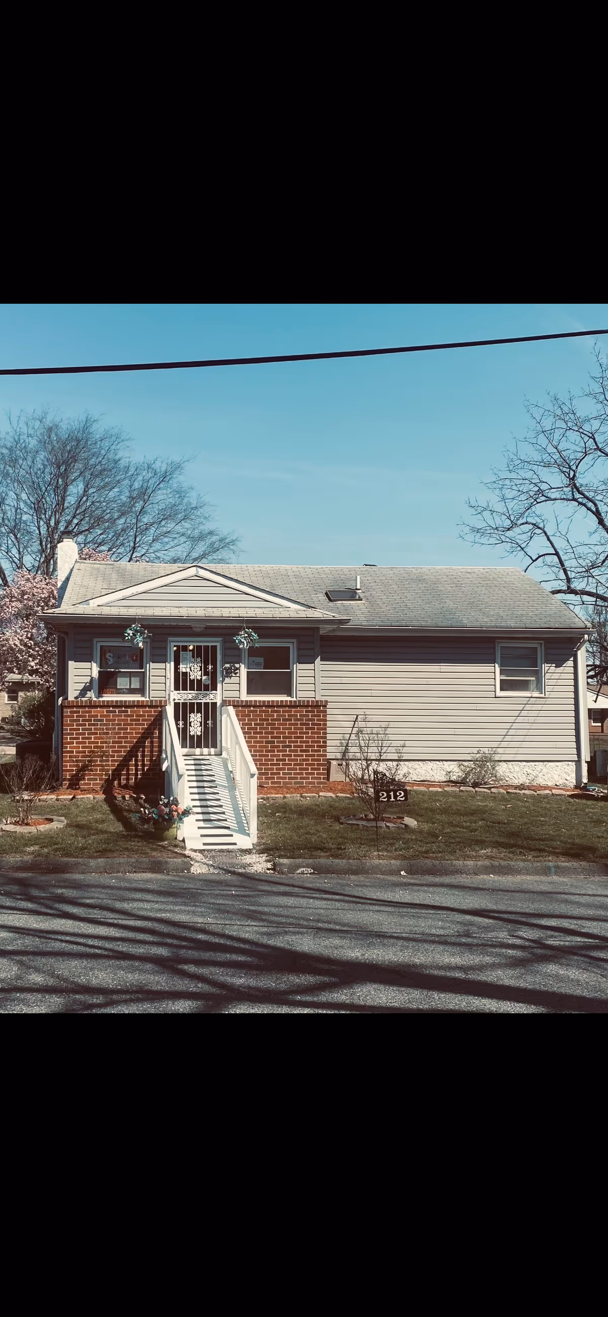 Single-story house with a small front porch and a ramp leading up to the entrance. The house has beige siding and a brick base around the porch area. There are a few small plants and a lawn in front, with a sign displaying the number 212. Trees without leaves are visible in the background under a clear blue sky.