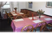 A dining room with two long tables covered with pink tablecloths, each set with placemats and napkins. Wooden chairs with curved backs surround the tables. There are flower arrangements in vases on the tables, and windows with sheer curtains in the background. Artwork is hung on the walls.