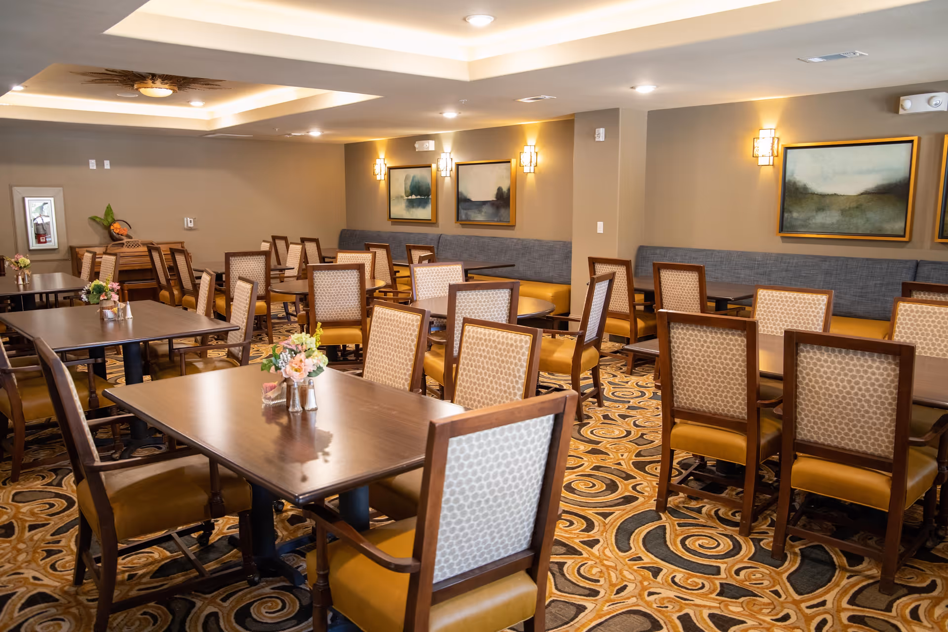 A well-lit dining room with multiple wooden tables and cushioned chairs arranged neatly. The room features a patterned carpet, wall-mounted lights, framed artwork on the walls, and a piano in the corner. Small floral centerpieces are placed on the tables.