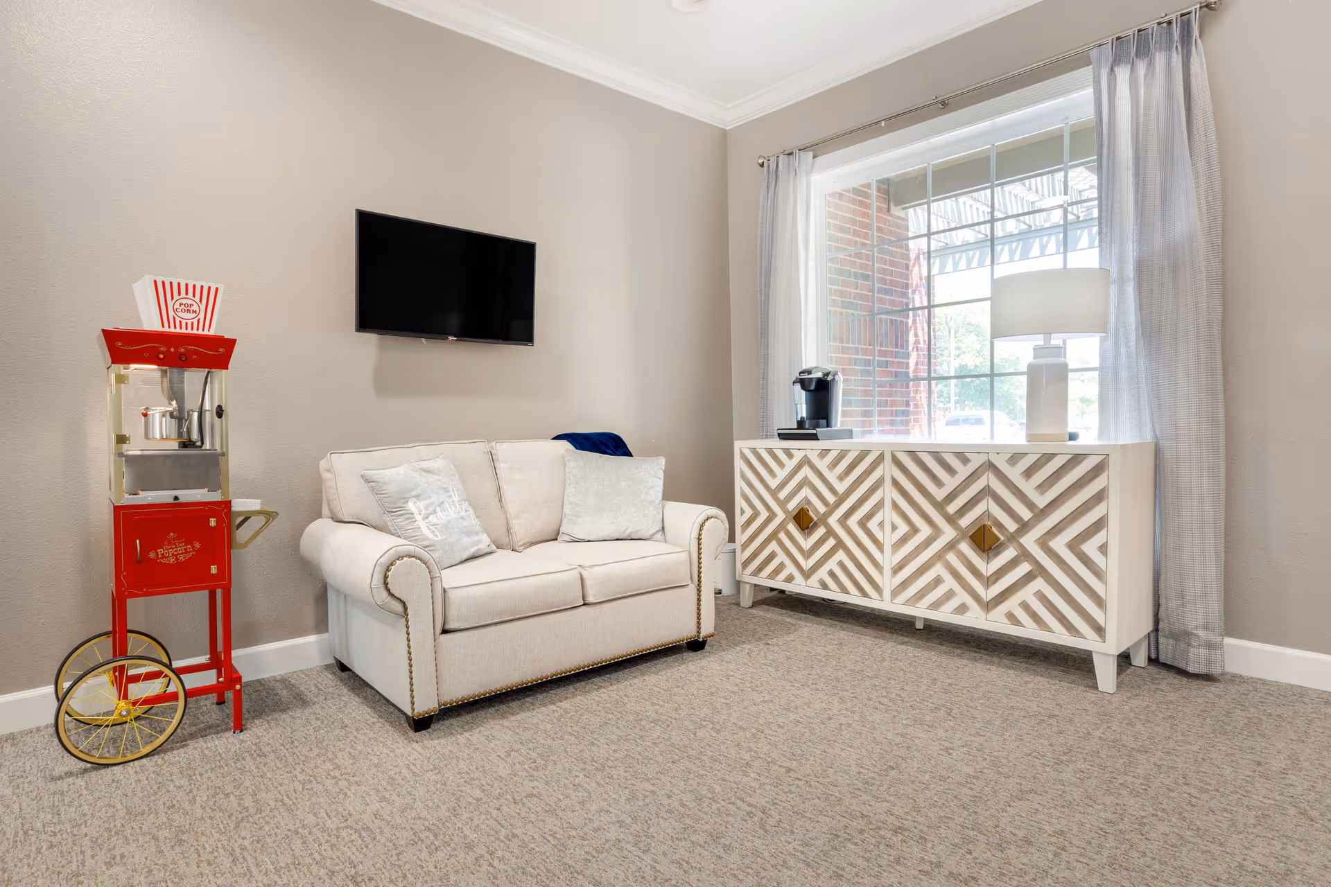 A cozy senior living room with a beige loveseat adorned with two pillows, a wall-mounted flat-screen TV, a vintage-style red popcorn machine on wheels, and a decorative cabinet with a geometric pattern under a large window with light curtains. A white table lamp and a coffee maker sit on top of the cabinet.