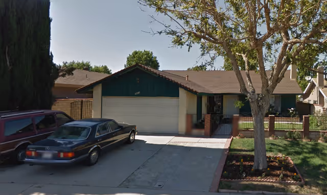 Single-story residential house with a two-car garage, a driveway with two parked cars, a front yard with a tree and flower bed, and a brick fence surrounding the yard.