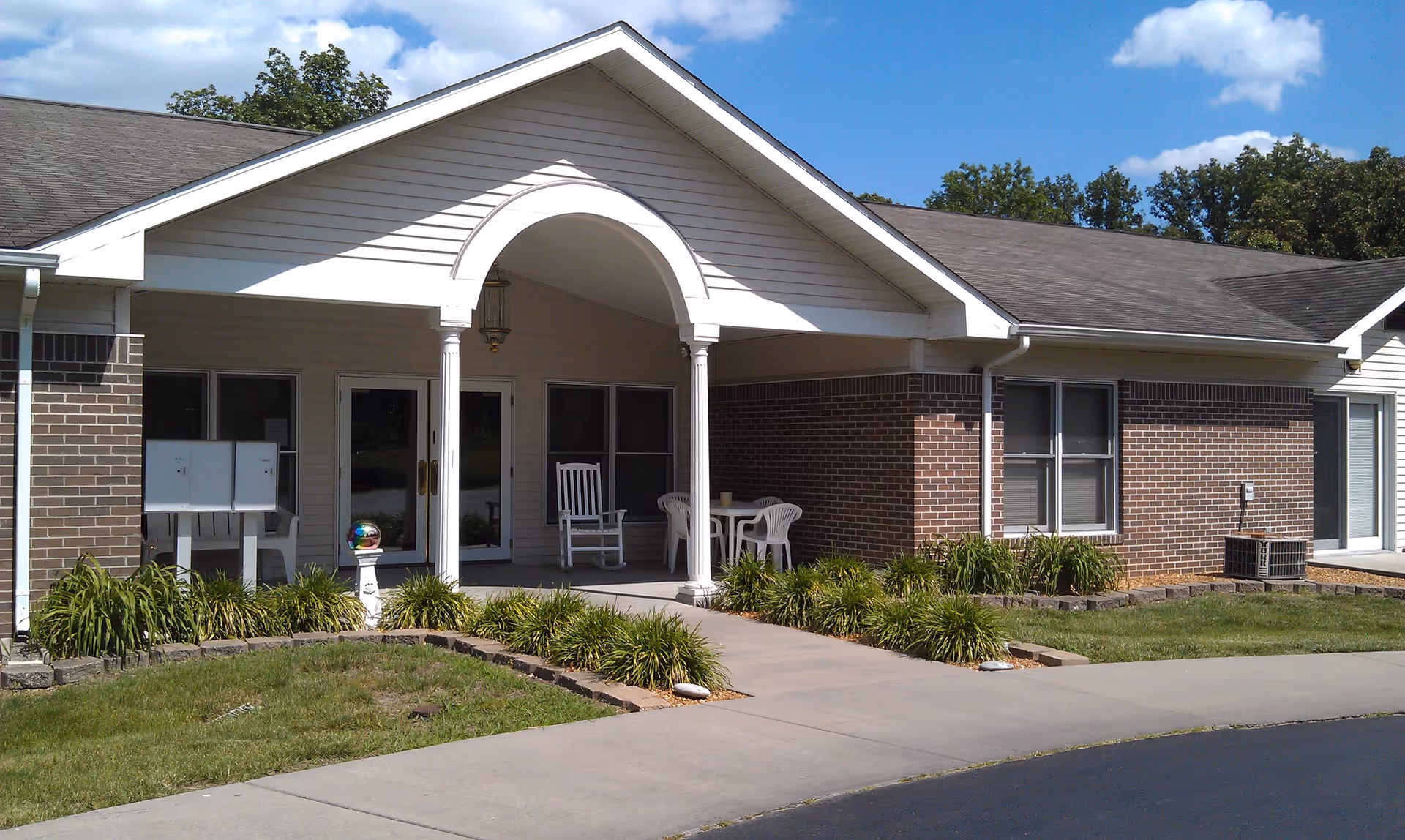 Exterior view of a single-story brick and siding building with a covered entrance supported by white columns. There is a white rocking chair and a small table with chairs on the porch. The building is surrounded by green grass and landscaping with bushes. The sky is blue with some clouds.