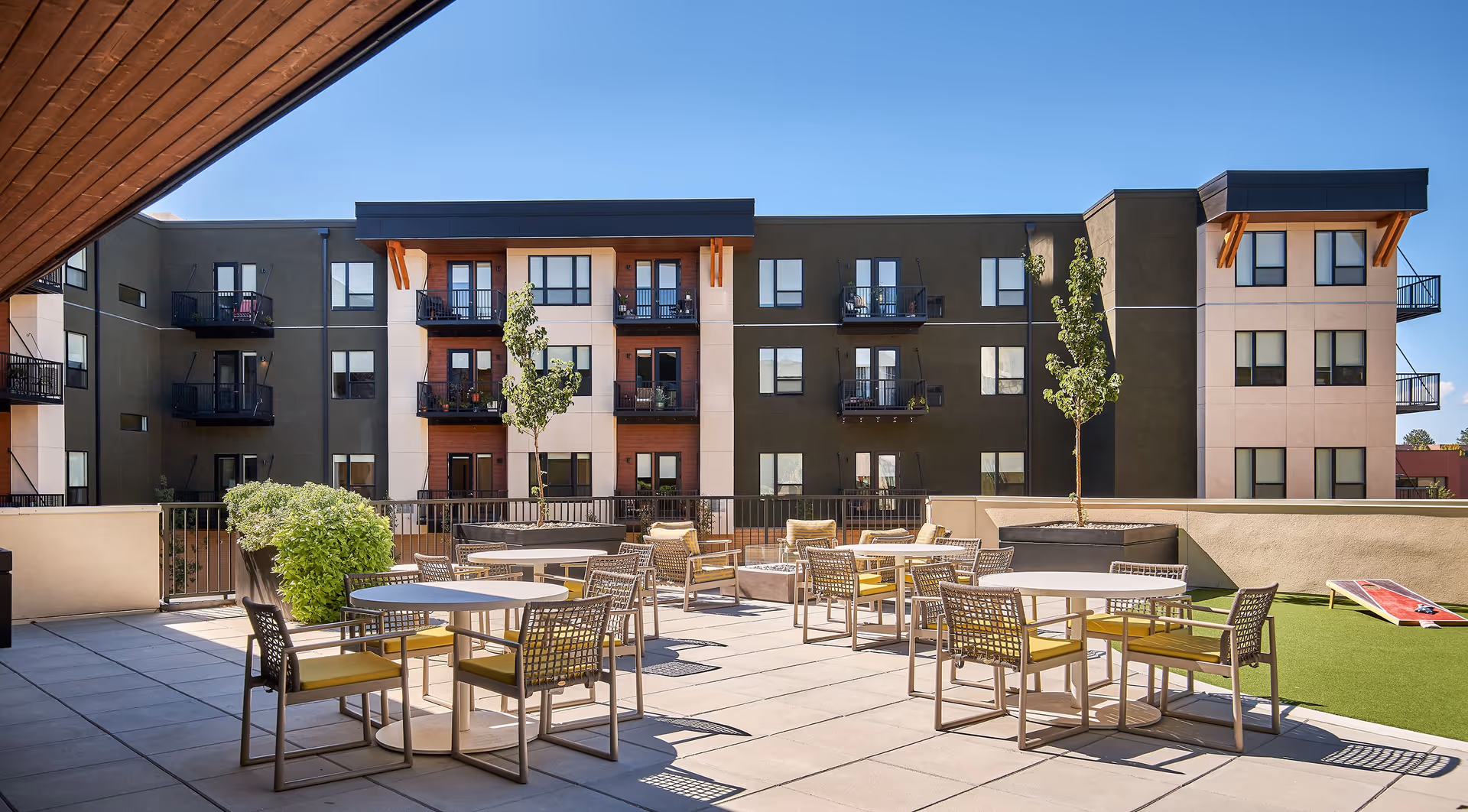 Rooftop outdoor patio with tables, chairs, planters and a grassy area in front of a multi-story residential building.