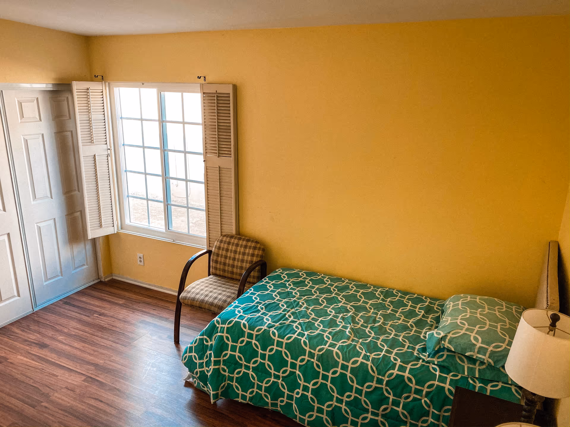 Small bedroom with a single bed in green patterned bedding, a chair by a window, and yellow walls.