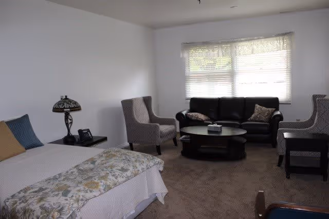 A senior living facility room with a bed on the left side covered with a floral bedspread and pillows. Next to the bed is a small table with a decorative lamp and a telephone. In the background, there is a seating area with a black leather sofa, two patterned armchairs, and a round coffee table. A large window with blinds lets in natural light.