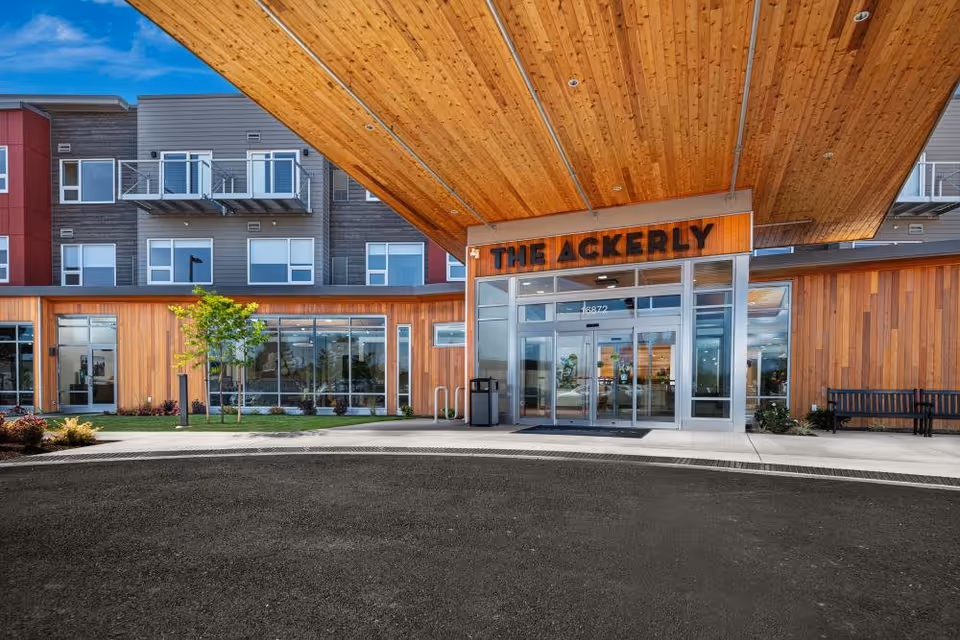 Exterior front entrance of a modern senior living facility named The Ackerly, featuring large glass doors, wooden paneling, and a covered driveway area with a bench and landscaping.