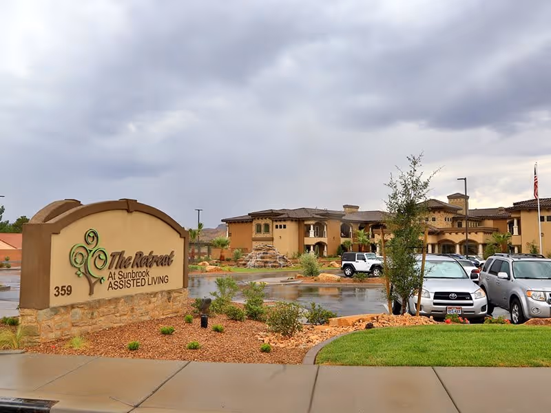 Exterior view of The Retreat at Sunbrook assisted living facility with a large sign in the foreground and the building complex, parking lot, and several cars in the background under a cloudy sky.
