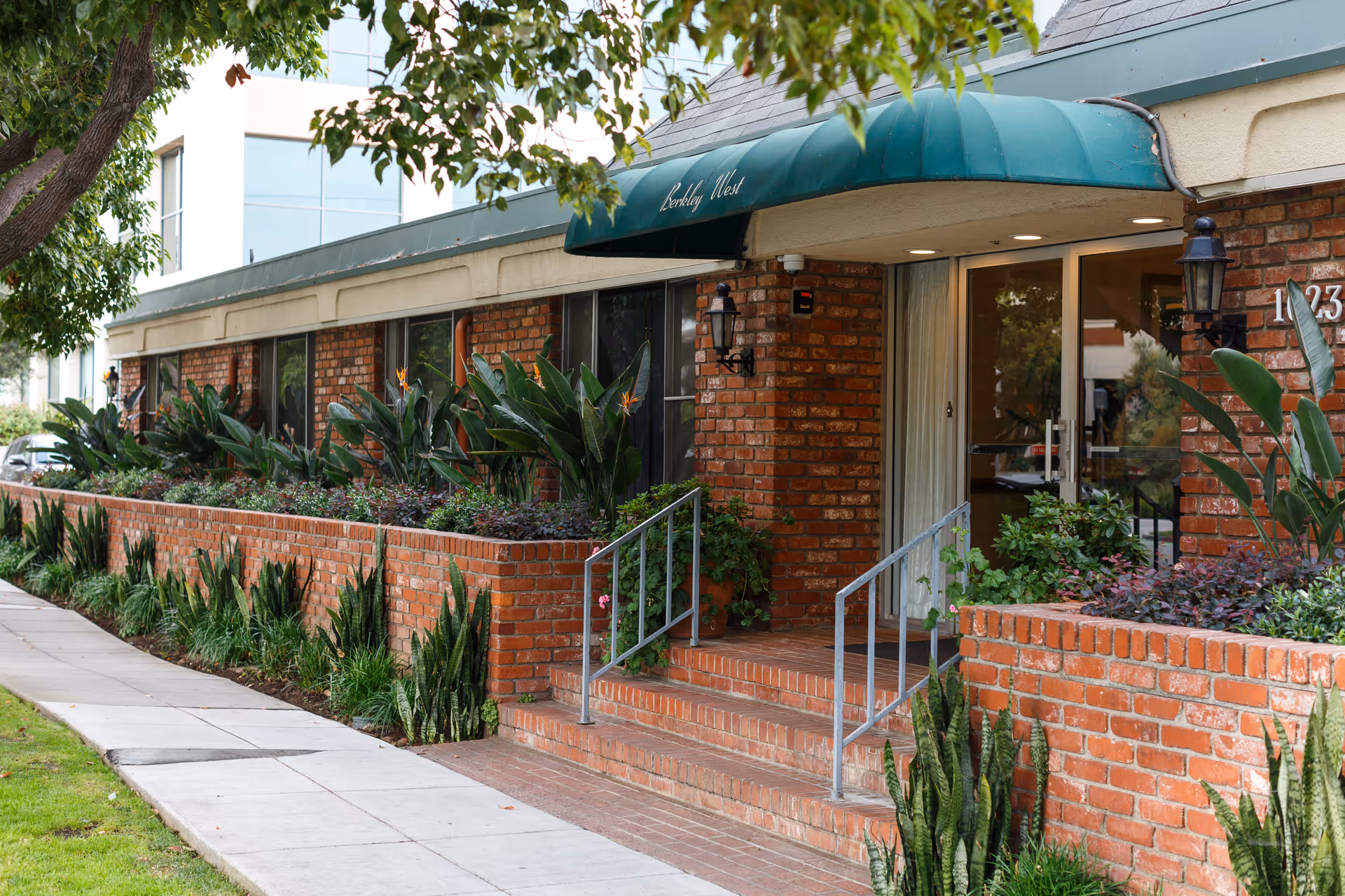 Exterior view of Berkley West Healthcare Center showing a brick building with a green awning above the entrance. There are plants and shrubs in brick planters along the sidewalk leading to the entrance.