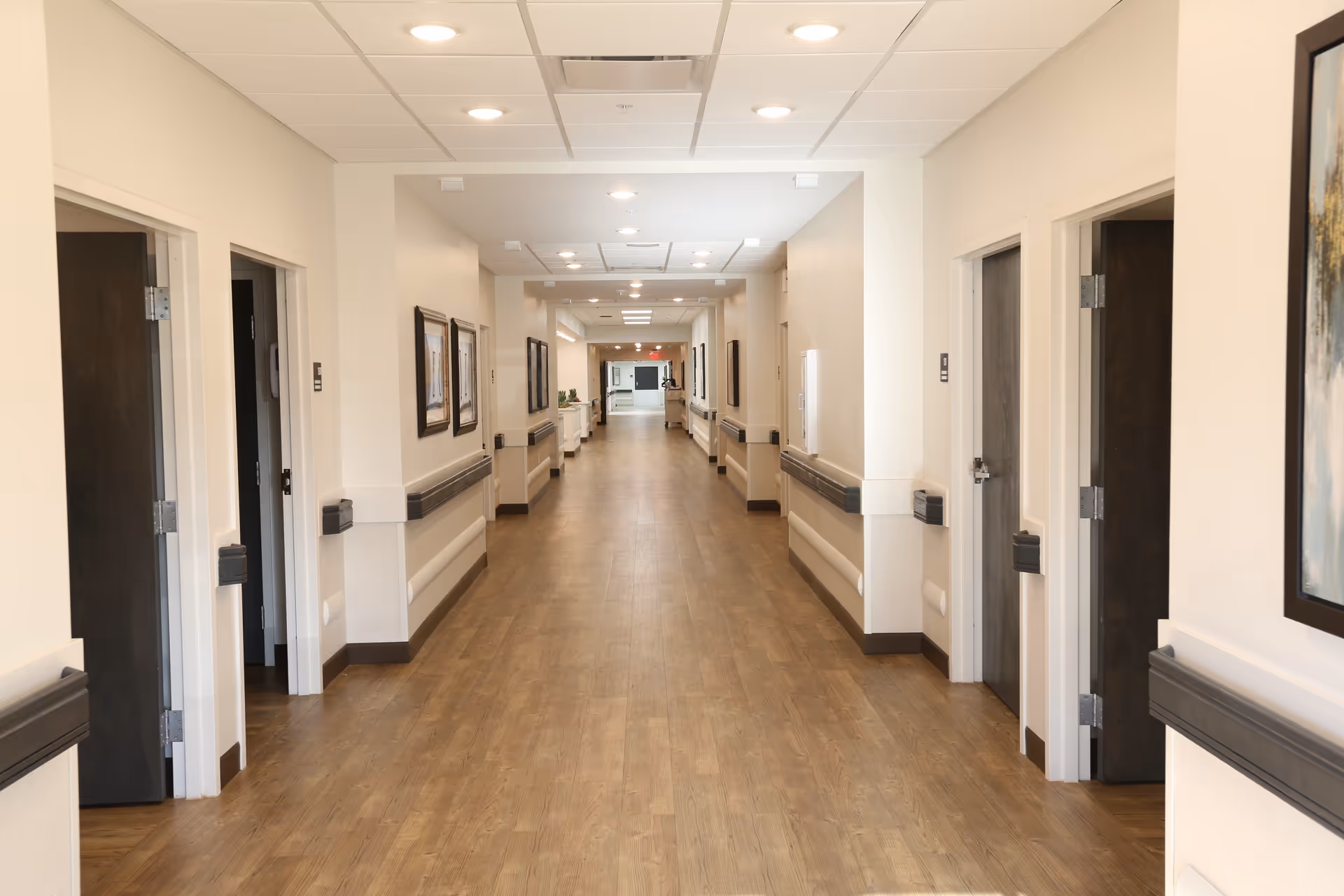 A long, well-lit hallway in a senior living facility with wooden flooring, beige walls, and multiple open doors on both sides. The hallway has handrails along the walls and framed artwork hanging on the walls.