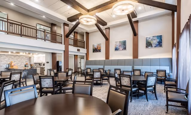 A spacious dining room with multiple dark wood tables and chairs arranged neatly. The room features high ceilings with exposed wooden beams, large round ceiling lights, and a mezzanine balcony. There are three framed artworks on the wall above a long cushioned bench. Large windows with curtains allow natural light to fill the space.