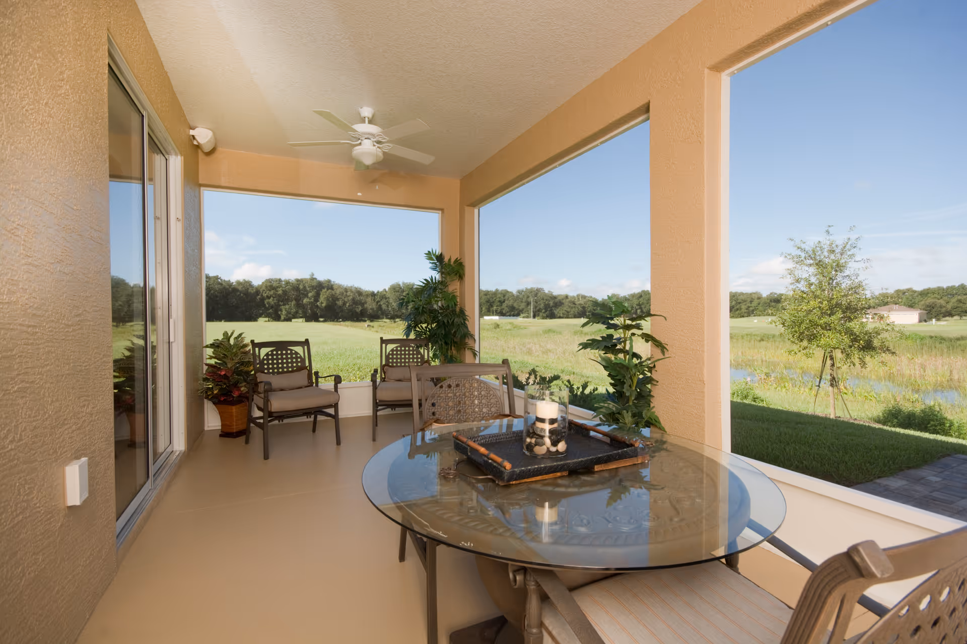 Covered outdoor patio area with a glass-top round table and cushioned chairs. The patio overlooks a green field with trees in the distance. There are potted plants placed around the seating area and a ceiling fan above.
