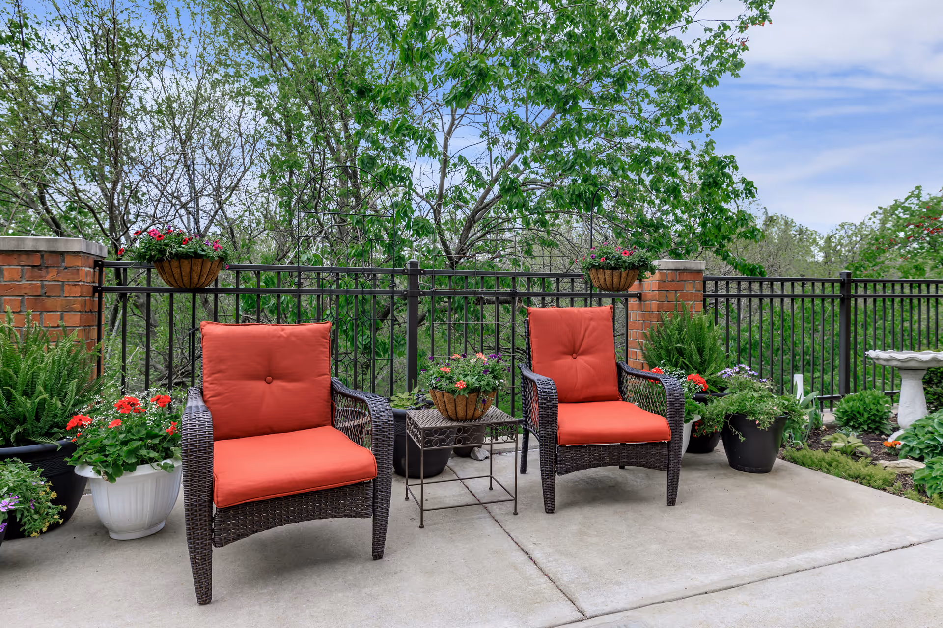 Two wicker patio chairs with red cushions and a small table on a concrete terrace surrounded by potted plants and trees.