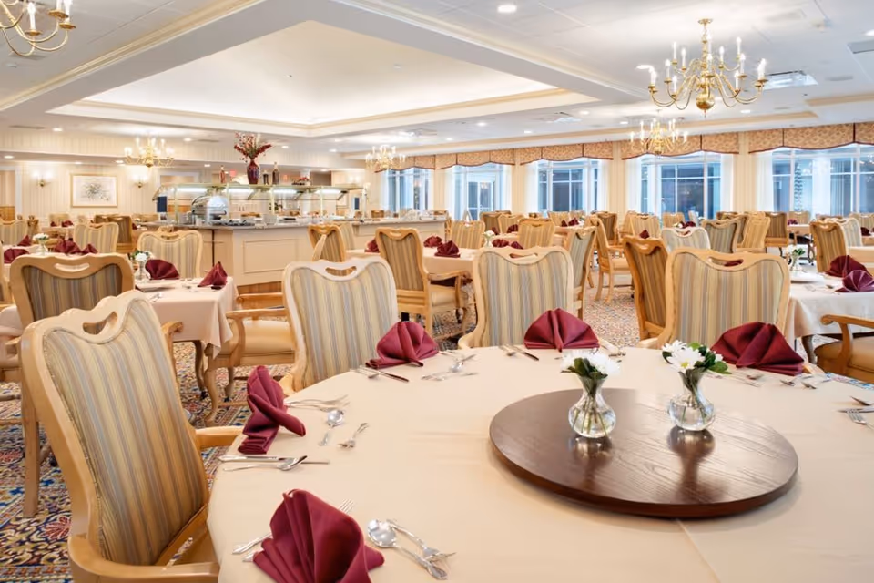 A spacious dining room with multiple round tables covered in beige tablecloths, each set with burgundy folded napkins, silverware, and small vases with white flowers. The room features striped upholstered chairs, chandeliers hanging from the ceiling, large windows with valances, and a buffet area in the background.