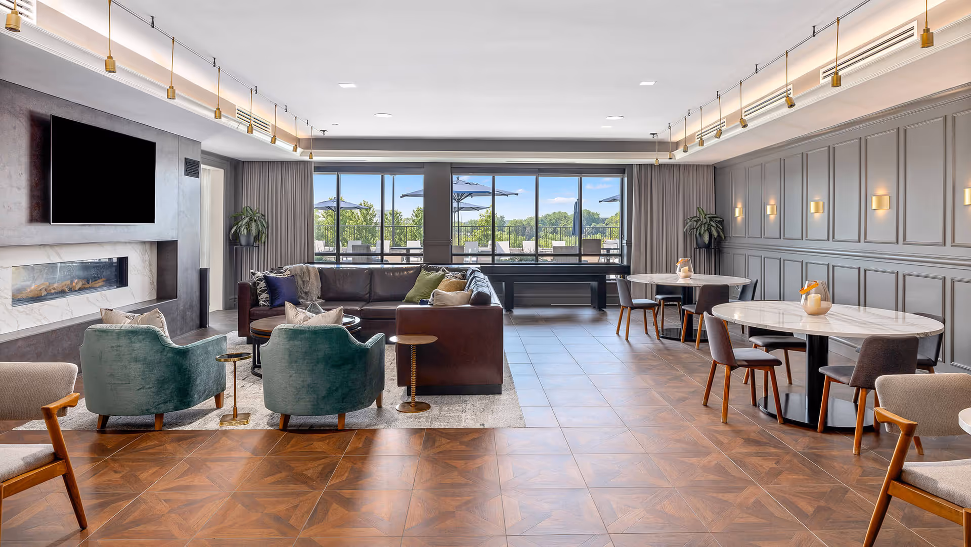A spacious and modern living room area with a large brown leather sectional sofa, two green armchairs, and a fireplace beneath a mounted flat-screen TV. The room features wood-patterned tile flooring, round tables with chairs on the right side, and large windows at the back showing an outdoor patio with umbrellas and greenery.
