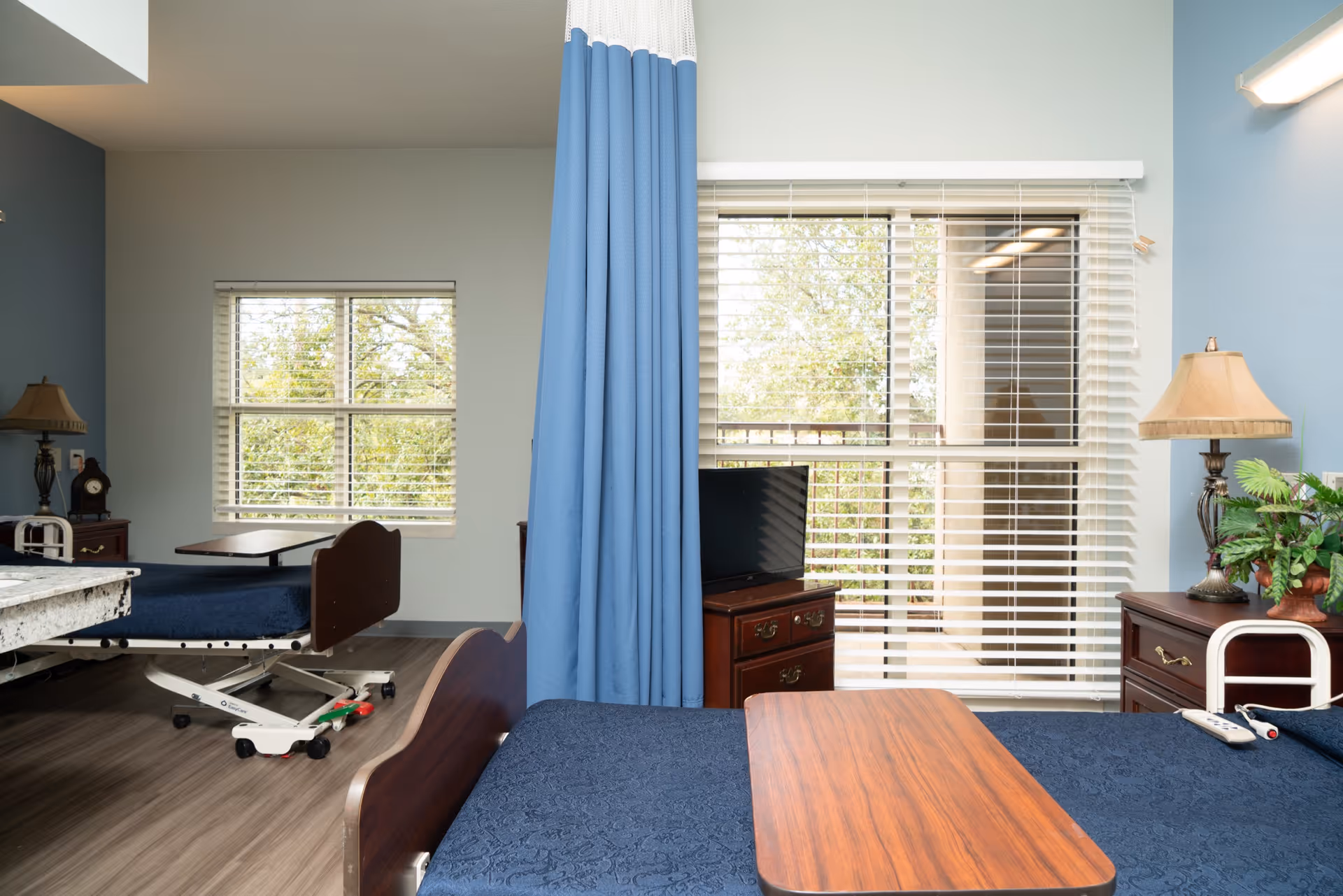 A healthcare facility room with two hospital beds separated by a blue privacy curtain. Each bed has a wooden overbed table. The room has large windows with white blinds, allowing natural light to enter. There are bedside tables with lamps and a small TV on one of the tables. The walls are painted light blue and the floor has wood-like vinyl flooring.