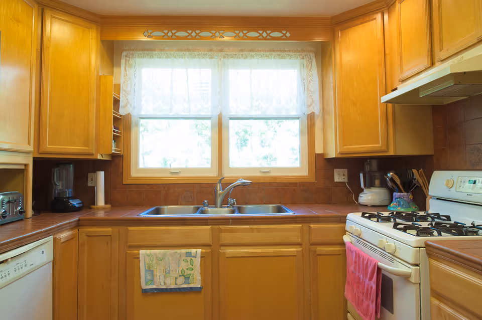 A kitchen with wooden cabinets, a double sink under a window with lace curtains, a white gas stove with a pink towel hanging on the handle, a blender, a toaster, and a food processor on the countertops.