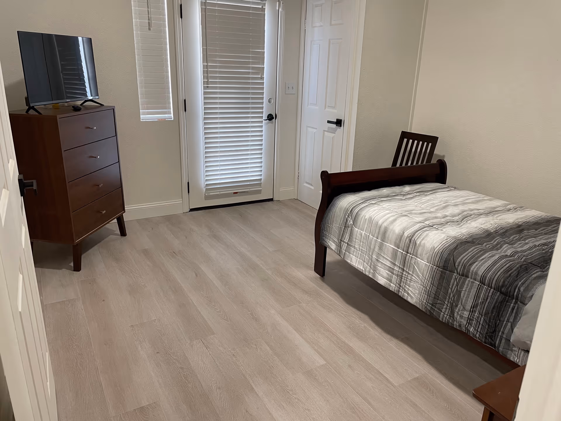 A simple bedroom with light wood flooring, a single bed with a gray and white striped comforter, a wooden chair, a wooden dresser with a flat-screen TV on top, a door with blinds, and a small window with blinds.