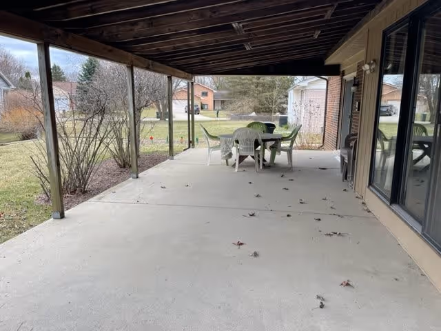 Covered outdoor patio area with a concrete floor, supported by wooden beams. There is a table with a green tablecloth and several white plastic chairs around it. The patio overlooks a grassy yard with some bushes and trees, and neighboring houses are visible in the background.